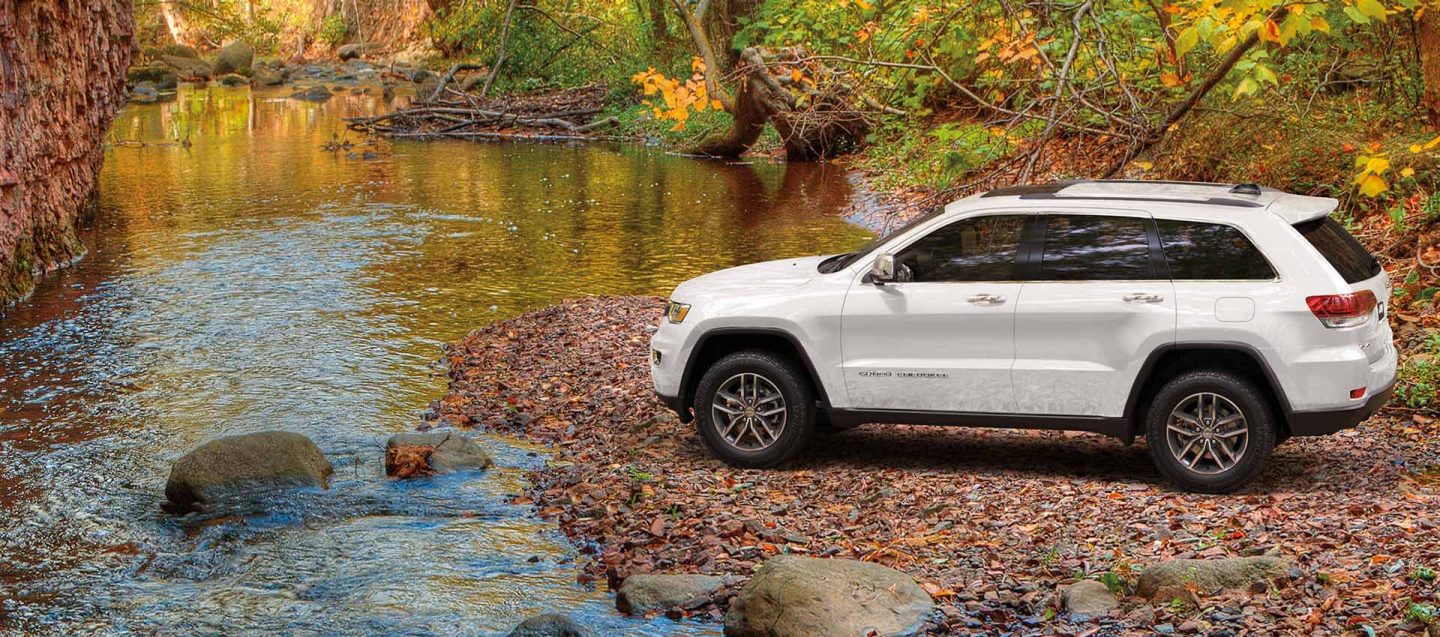 A white 2020 Jeep&reg; Grand Cherokee Limited parked facing a stream with trees and a rock wall in the background.